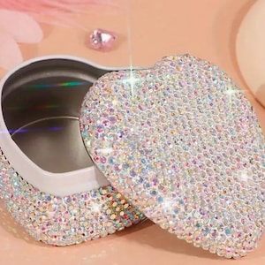 May include: A heart-shaped trinket box covered in iridescent rhinestones. The box is open, revealing a silver interior.