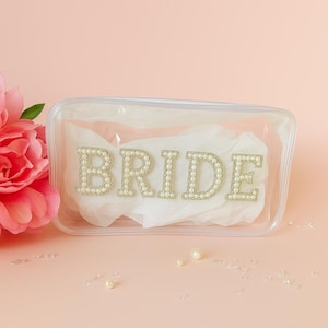 May include: A clear, rectangular cosmetic bag with the word "BRIDE" spelled out in gold-colored letters, each outlined with small, round, pearl-like beads. The bag contains white tissue paper and sits on a pink surface.