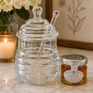 May include: A clear glass honey pot with a matching lid and a glass dipper, featuring horizontal ribbed detailing and Hebrew text etched on the front. Beside it sits a small glass jar of Tiptree Pure Honey with a silver lid and a label stating 28g 1oz.