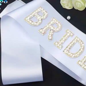 May include: White satin sash with the word "BRIDE" spelled out in rhinestones. The sash is perfect for a wedding or bridal shower.