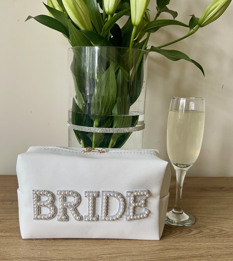 May include: White zippered makeup bag with the word "BRIDE" spelled out in pearl-like embellishments.