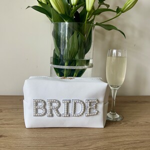 May include: White zippered makeup bag with the word "BRIDE" spelled out in pearl-like embellishments. The bag is sitting on a wooden surface with a glass of champagne and a vase of white lilies in the background.