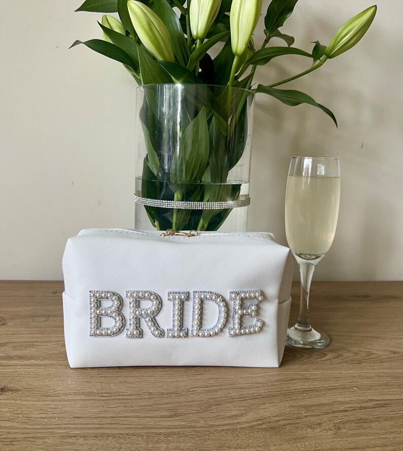 May include: White zippered makeup bag with the word "BRIDE" spelled out in pearl-like embellishments. The bag is sitting on a wooden surface with a glass of champagne and a vase of white lilies in the background.