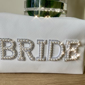 May include: White cosmetic bag with the word "BRIDE" spelled out in faux pearls.