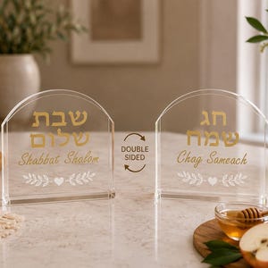 Könnte beinhalten: Ein doppelseitiges, bogenförmiges Schild aus klarem Acryl mit goldenem hebräischem und englischem Text. Eine Seite zeigt Shabbat Shalom mit einem Blatt- und Herzmotiv, die andere Seite zeigt Chag Sameach mit einem passenden Blatt- und Herzdesign.