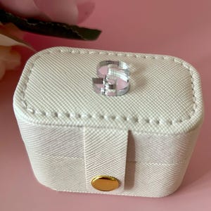 May include: A white, rectangular jewelry box with a gold button and a clear acrylic letter 'S' on the top.