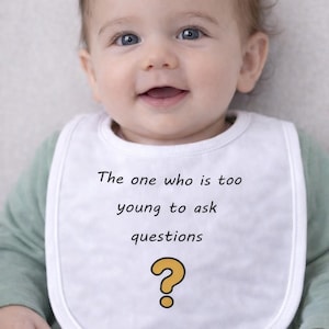 May include: A baby wearing a white bib with the text "The one who is too young to ask questions" and a yellow question mark. The baby smiles, showing blue eyes. The baby wears a green long-sleeved top.