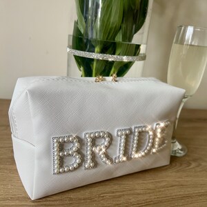 May include: White cosmetic bag with the word "BRIDE" spelled out in faux pearls. The bag is on a wooden surface.