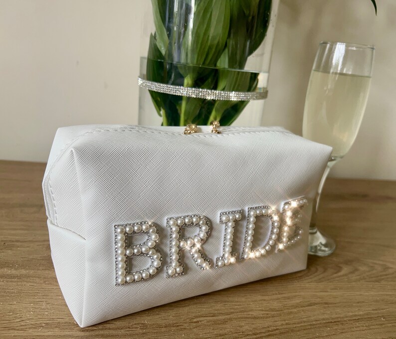 May include: White cosmetic bag with the word "BRIDE" spelled out in faux pearls. The bag is on a wooden surface.