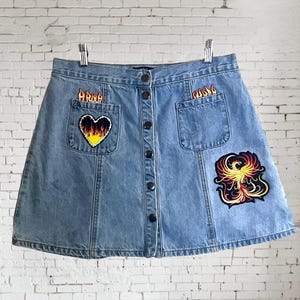 Könnte beinhalten: Hellblauer Jeansrock mit Knopfleiste und zwei Vordertaschen. Der Rock hat gestickte Aufnäher mit Flammen, einem Herzen mit Flammen und einem Phönix. Der Rock hängt an einem Metallbügel vor einer weißen Backsteinmauer.