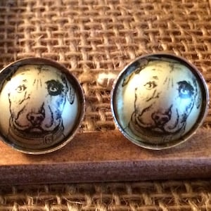 May include: A pair of round silver-toned stud earrings. Each earring features a detailed black and white illustration of a dog's face. The earrings are set against a textured, woven background.