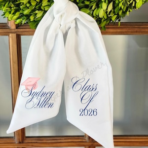 May include: White graduation sash with the text "Sydney Allen" and "Class Of 2026" in navy blue script. A pink graduation cap graphic is also present. The sash is tied in a knot and hangs in front of a green wreath.