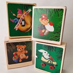 May include: Set of four Christmas ornaments in boxes. The ornaments are a gnome in a fortune cookie, a gnome playing a drum, a teddy bear with a rattle, and a bunny on roller skates. The boxes have green backgrounds with images of the ornaments and text that says "Sixth in a collectible series" and "Look for series verification on this ornament."