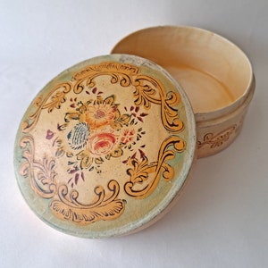 May include: An open, round, vintage box with a floral design on the lid. The lid features a cream-colored background with a floral arrangement in the center, surrounded by gold scrollwork. The box is cream-colored.