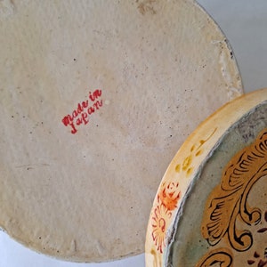 May include: Close-up of a vintage, round, ceramic container with a lid. The lid is off, revealing the interior and the words "made in Japan" in red. The rim of the container is decorated with floral and scroll designs.