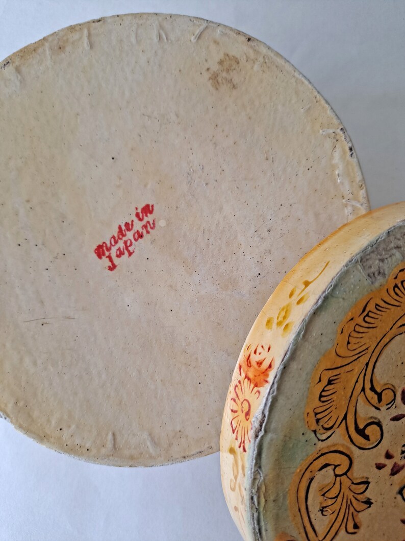 May include: Close-up of a vintage, round, ceramic container with a lid. The lid is off, revealing the interior and the words "made in Japan" in red. The rim of the container is decorated with floral and scroll designs.