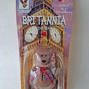 May include: A brown beanie baby bear wearing a red ribbon and a patch with the Union Jack flag. The bear is packaged in a plastic bag with a Ty tag and a background image of Big Ben. The tag reads "Britannia The Bear" and "Collect All 4! While supplies last."