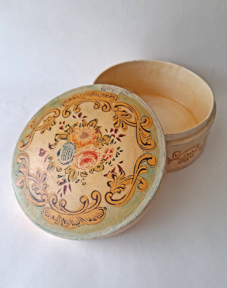 May include: An open, round, vintage box with a floral design on the lid. The lid features a cream-colored background with a floral arrangement in the center, surrounded by gold scrollwork. The box is cream-colored.