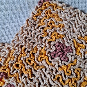 May include: A crocheted textile featuring a dense, wavy, and textured pattern. The design incorporates a repeating color palette of beige, golden yellow, and muted mauve, arranged in interconnected, undulating lines that create a complex surface.