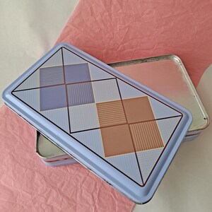 May include: A rectangular, light blue tin with a geometric design of squares in lavender, tan, and light blue. The tin is open, revealing a silver interior, and rests on a pink paper surface.