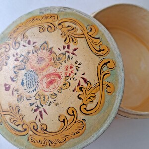 May include: Round, vintage trinket box with a floral design on the lid. The lid features a cream-colored background with a floral bouquet in the center and gold scrollwork around the edge. The box is open, revealing the interior.