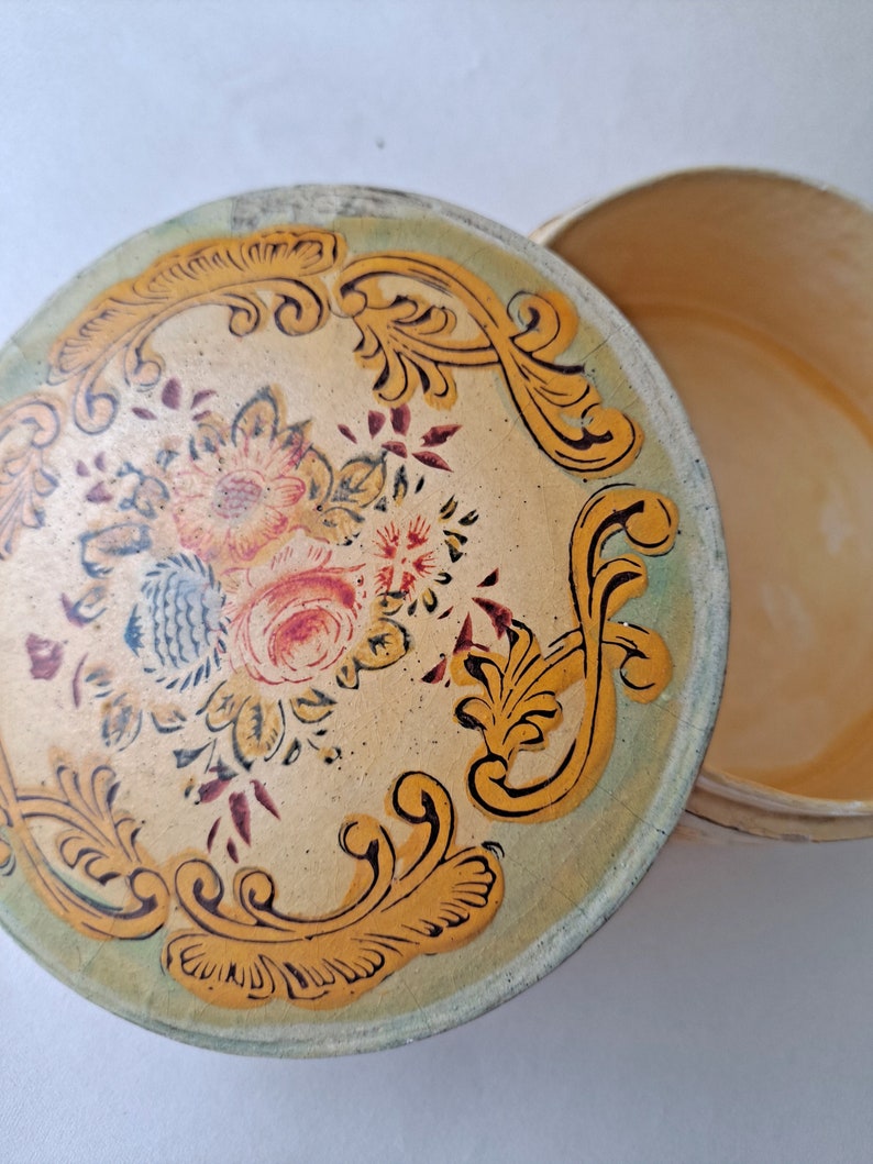 May include: Round, vintage trinket box with a floral design on the lid. The lid features a cream-colored background with a floral bouquet in the center and gold scrollwork around the edge. The box is open, revealing the interior.