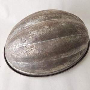 May include: A vintage metal mold for baking a loaf of bread in the shape of a melon. The mold is made of silver-colored metal and has a slightly worn appearance.