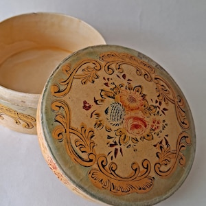 May include: An open, round, vintage box with a floral design on the lid. The lid features a central bouquet of red, yellow, and orange flowers surrounded by ornate, gold-colored scrollwork on a beige background. The box is beige with a green rim.