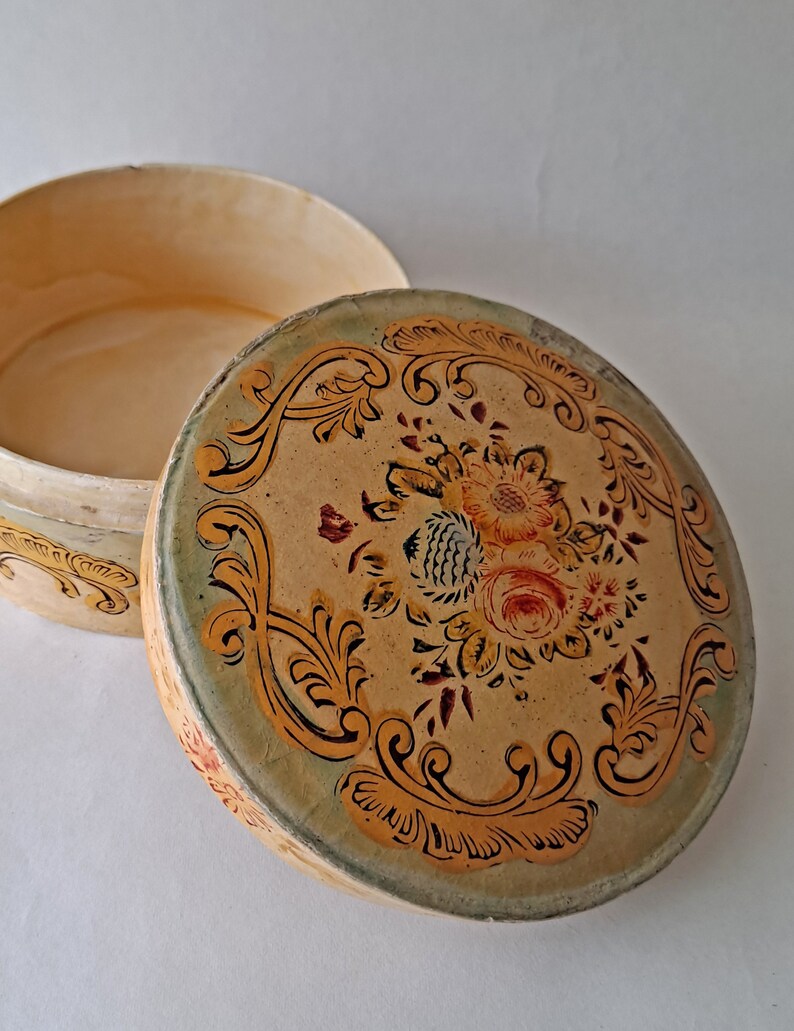 May include: An open, round, vintage box with a floral design on the lid. The lid features a central bouquet of red, yellow, and orange flowers surrounded by ornate, gold-colored scrollwork on a beige background. The box is beige with a green rim.