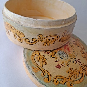 May include: An open, oval-shaped, antique box with a lid. The box is painted in cream and yellow tones, with gold and black floral scrollwork. The lid features a floral design with red and blue accents. The box is likely vintage.