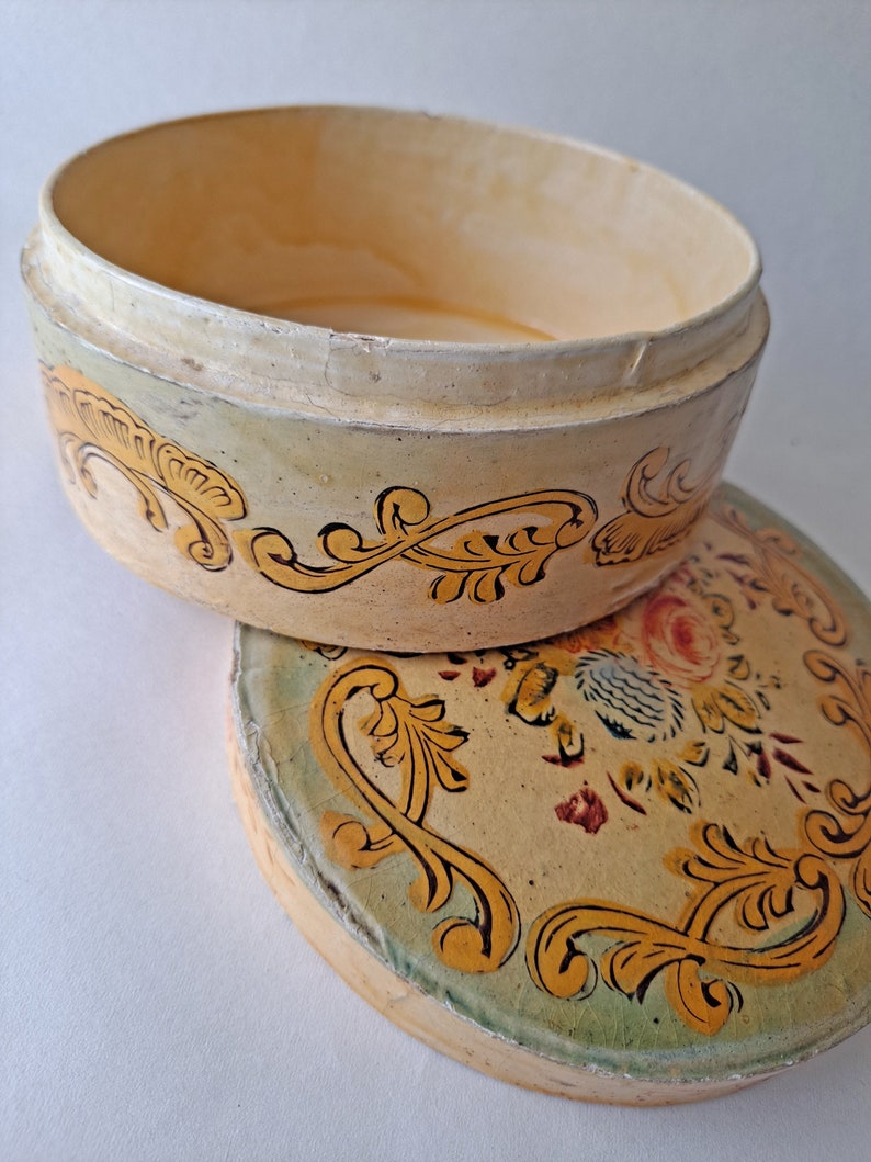 May include: An open, oval-shaped, antique box with a lid. The box is painted in cream and yellow tones, with gold and black floral scrollwork. The lid features a floral design with red and blue accents. The box is likely vintage.