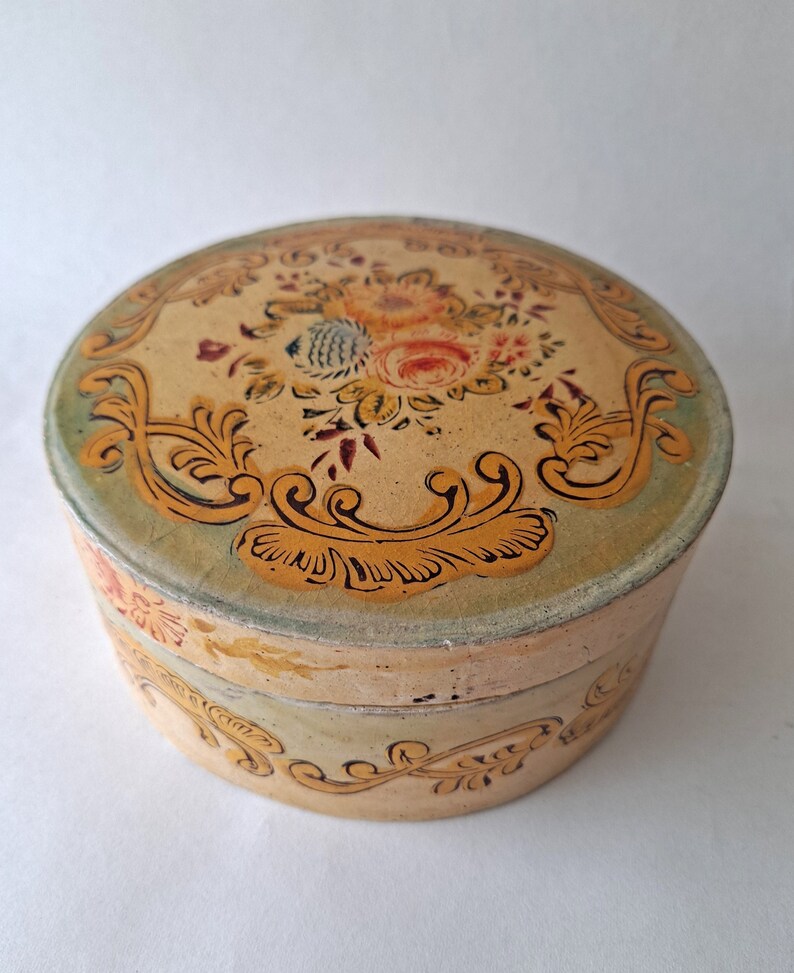 May include: A round, antique-style box with a floral design on the lid. The box is painted in cream, with a green border and gold-colored scrollwork. The lid features a bouquet of flowers in shades of pink, yellow, and blue.