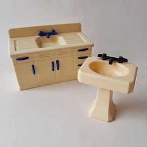 May include: A set of two white plastic dollhouse bathroom sinks. One sink is a pedestal sink with a black faucet. The other sink is a countertop sink with a blue faucet and a cabinet with blue handles.