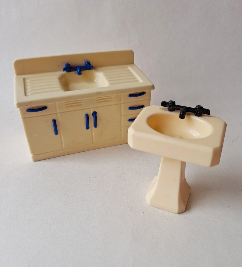May include: A set of two white plastic dollhouse bathroom sinks. One sink is a pedestal sink with a black faucet. The other sink is a countertop sink with a blue faucet and a cabinet with blue handles.