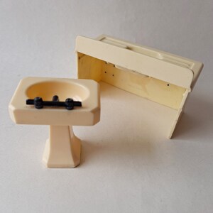 May include: A white plastic dollhouse bathroom sink and vanity set. The sink has a white pedestal and a black faucet. The vanity is a simple rectangular shape with a flat top.