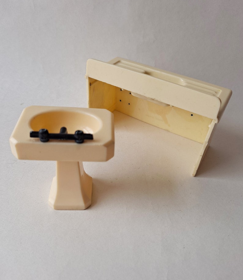 May include: A white plastic dollhouse bathroom sink and vanity set. The sink has a white pedestal and a black faucet. The vanity is a simple rectangular shape with a flat top.