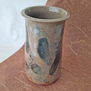 May include: A handmade ceramic vase with a cylindrical shape and a slightly flared rim. The vase features a mottled, earthy color palette of tan, brown, and dark blue, with abstract brushstroke-like patterns. The vase is suitable for displaying flowers or as a decorative item.