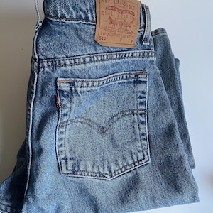 May include: Blue denim jeans featuring a tan leather patch on the waistband with the text Levi Strauss & Co. Original Riveted Quality Clothing Every Garment Guaranteed 512. A small red tab is visible on the edge of the back pocket.