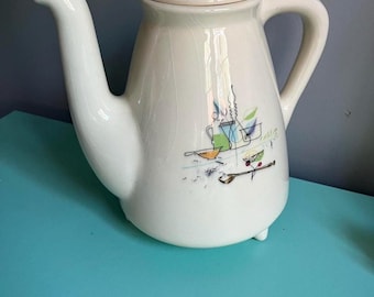 May include: White ceramic teapot with a stylized design of kitchen utensils and a bowl of fruit. The teapot has a lid and a handle.
