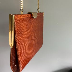 May include: A rectangular handbag with a textured, rust-colored surface featuring a fine, scale-like pattern. The bag is framed with polished gold-tone metal edges and a matching gold-tone clasp closure, suspended by a thin gold-tone chain strap.