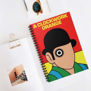 May include: A spiral bound notebook with a red, yellow and black cover featuring a minimalist illustration of Alex from the film "A Clockwork Orange". The title "A Clockwork Orange" is printed in yellow and black lettering on the cover.