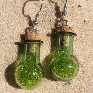 Puede incluir: Un par de pendientes únicos con pequeñas botellas de vidrio verde llenas de cuentas verdes. Las botellas están selladas con corchos y suspendidas de ganchos plateados. Los pendientes están sobre un fondo arenoso.