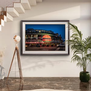 May include: Framed print of Wrigley Field at dusk, home of the Chicago Cubs. The image shows the stadium's exterior with illuminated signage and lights. The print is displayed in a room with a modern floor lamp and a potted plant.
