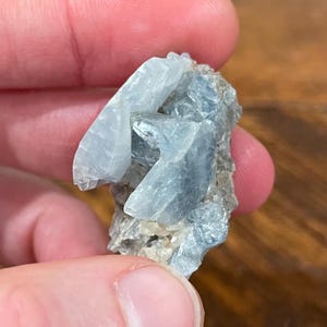 May include: A close-up of a light blue celestite crystal cluster. The crystal has a layered appearance with sharp, angular formations. The base of the crystal is a mix of gray and brown hues. The crystal is held in a person's hand.