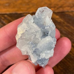 May include: A cluster of light blue celestite crystals. The crystals have a translucent appearance with a mix of light and darker blue hues. The mineral is held in a hand, with the background out of focus.