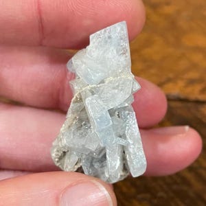 May include: A cluster of pale blue crystal formations. The crystals have a translucent quality and are arranged in a layered, geometric pattern. The mineral specimen is held between fingers, showcasing its natural beauty.