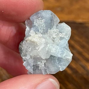 May include: A light blue celestite crystal cluster. The crystal has a rough, textured surface with small, clear crystal formations. The color is a pale, almost translucent blue, with some areas appearing white. The crystal is held between two fingers.