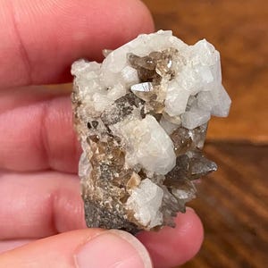 May include: A cluster of quartz crystals, featuring a mix of clear, white, and smoky brown hues. The crystals are tightly packed together, creating a textured, natural appearance. The specimen is held in a hand, showcasing its size and detail.