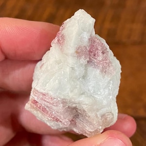 May include: A raw, uncut gemstone with a crystalline structure. The stone features a pale, off-white base color with streaks and patches of pink and red throughout. The mineral is held in a hand.
