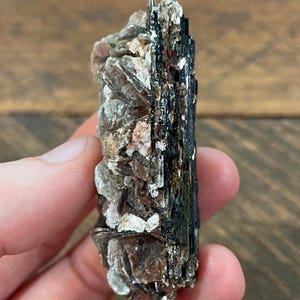 May include: A natural mineral specimen with a rough texture. One side displays a black, crystalline formation, while the other shows a mix of grey, brown, and pinkish-white flakes. The mineral is held in a hand against a wooden background.
